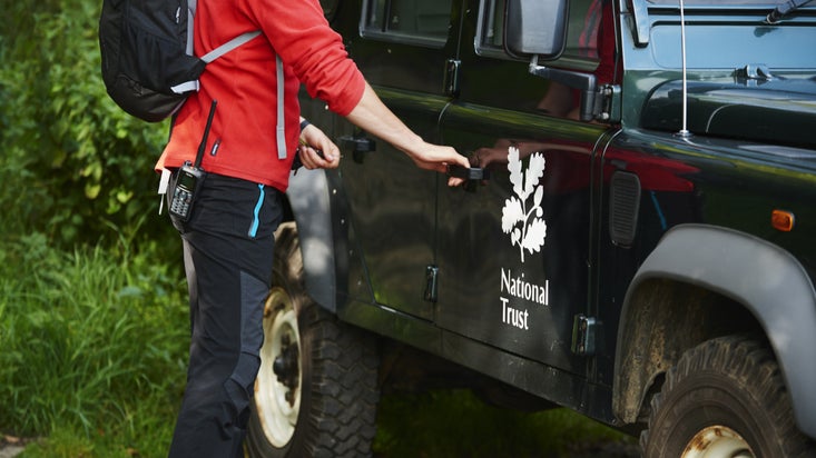 A close-up of a ranger with their torso and legs visible, wearing a red fleece and standing next to a National Trust branded vehicle